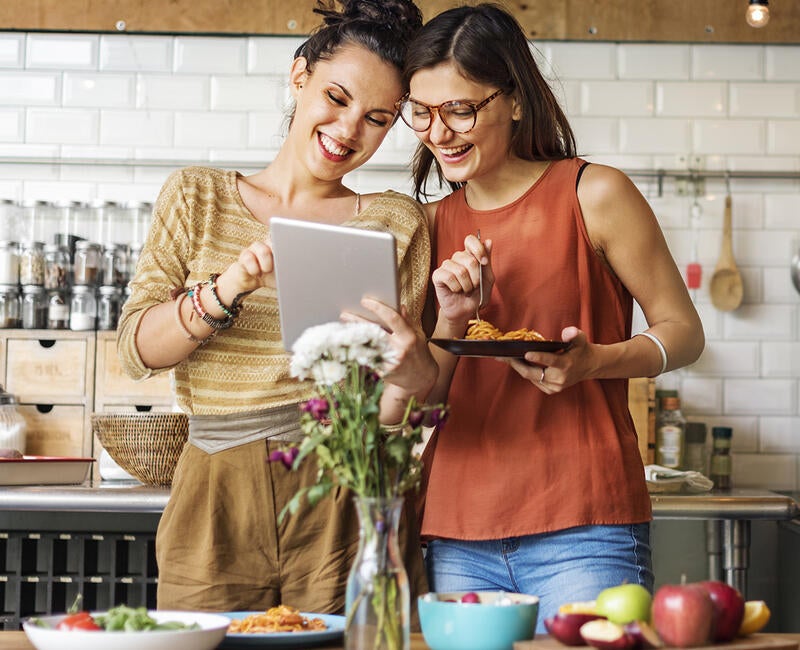 Two smiling young women use a tablet in a modern kitchen.
