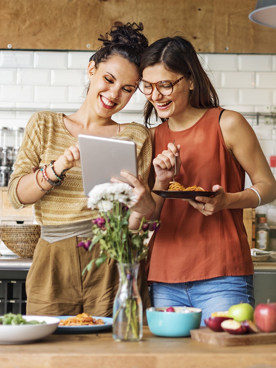 Two smiling young women use a tablet in a modern kitchen.