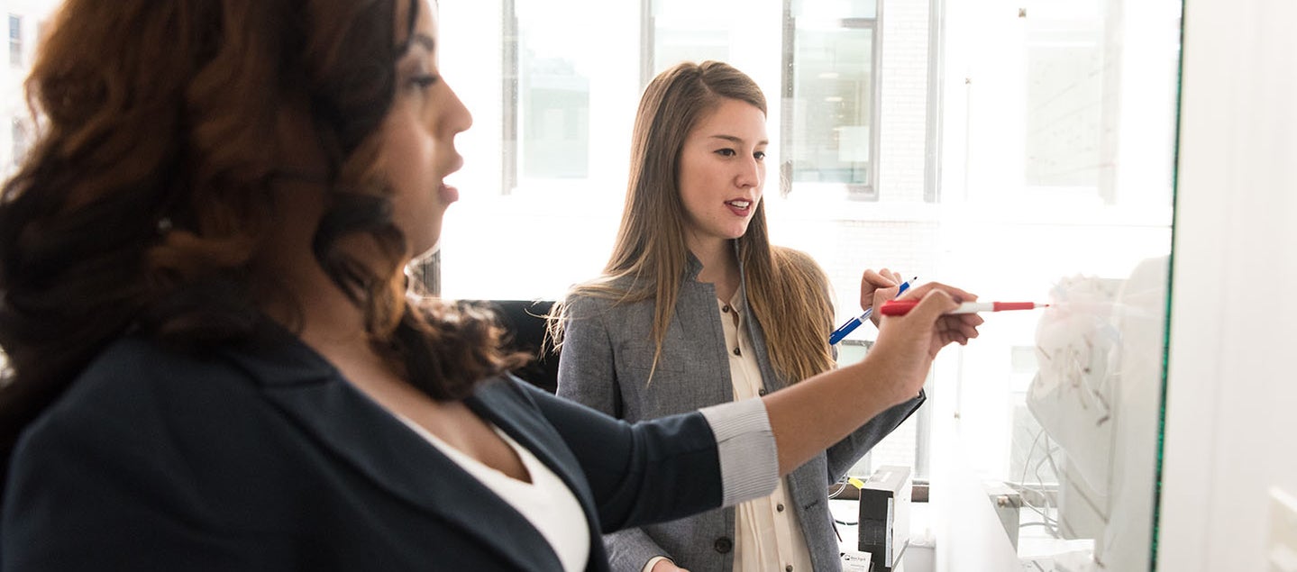 Women in business attire have a meeting around a whiteboard.