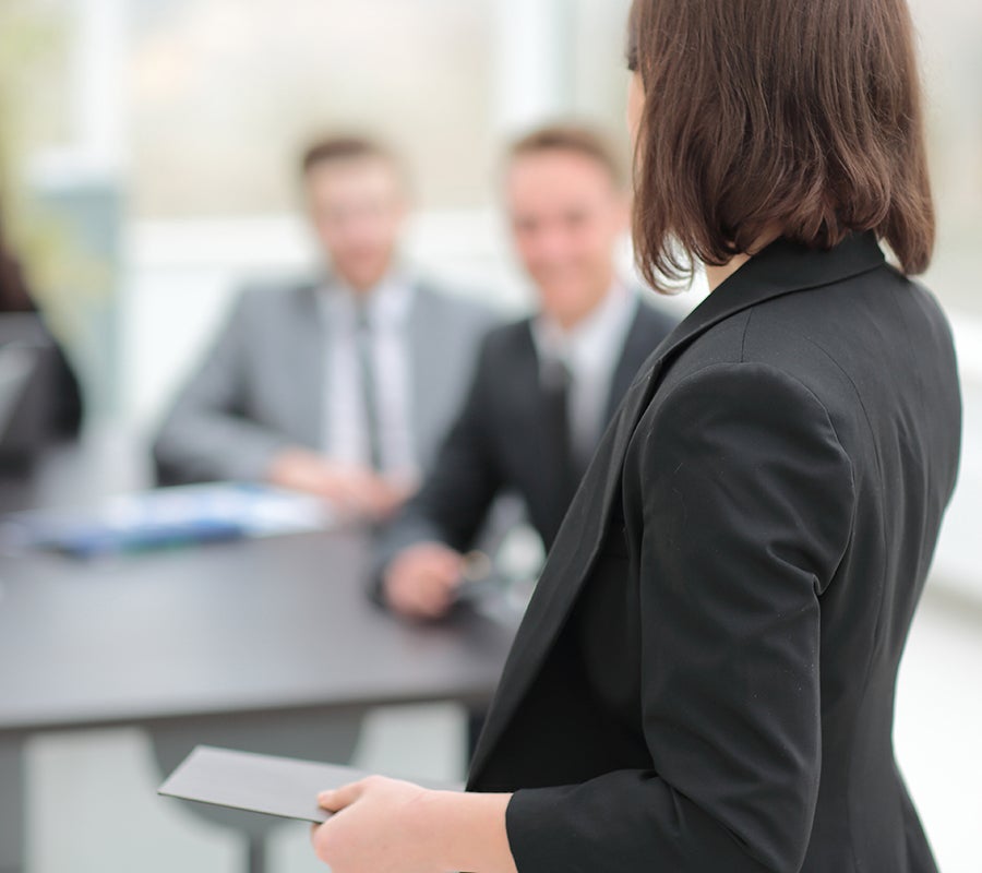 A woman in a blazer presents to a room full of businessmen.